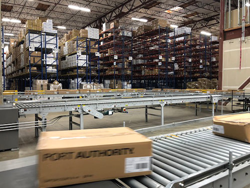 Background shows warehouse with very high pallet racking. Foreground shows serpentine conveyor with boxes being moved at high speed.